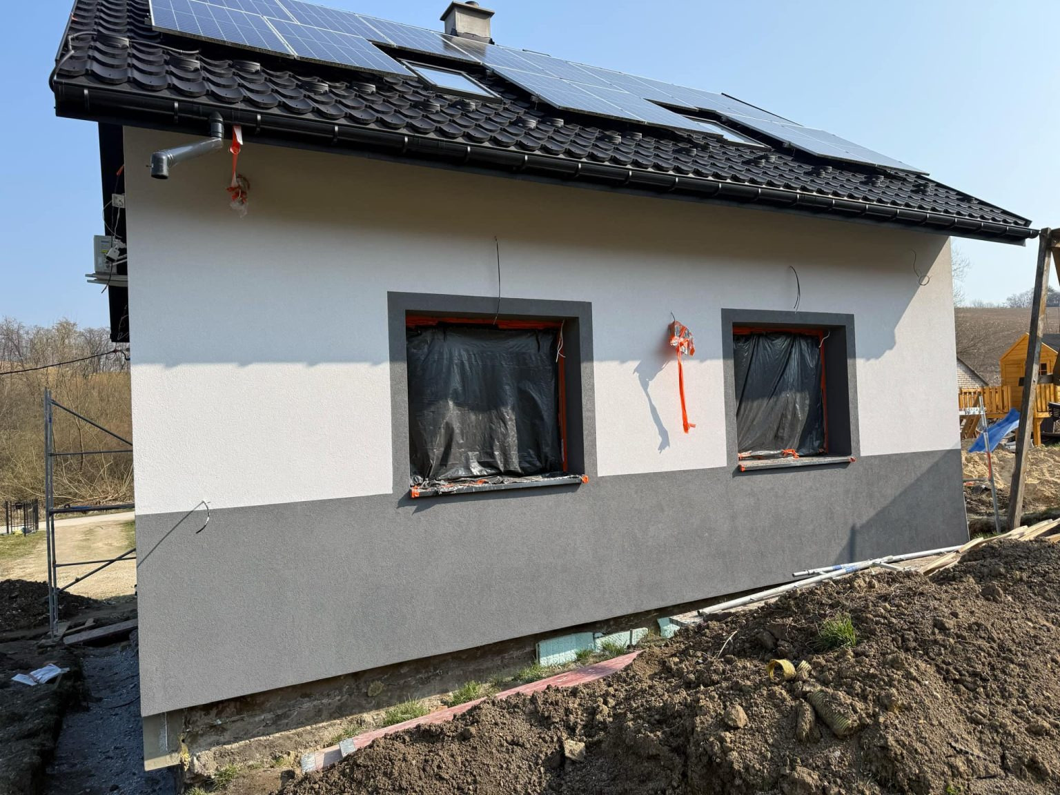 Ocieplona elewacja domu z panelami słonecznymi na dachu. Okna zabezpieczone folią. Widoczny fragment wykopu i ziemi. Dwukolorowa elewacja: biała góra, szary dół.