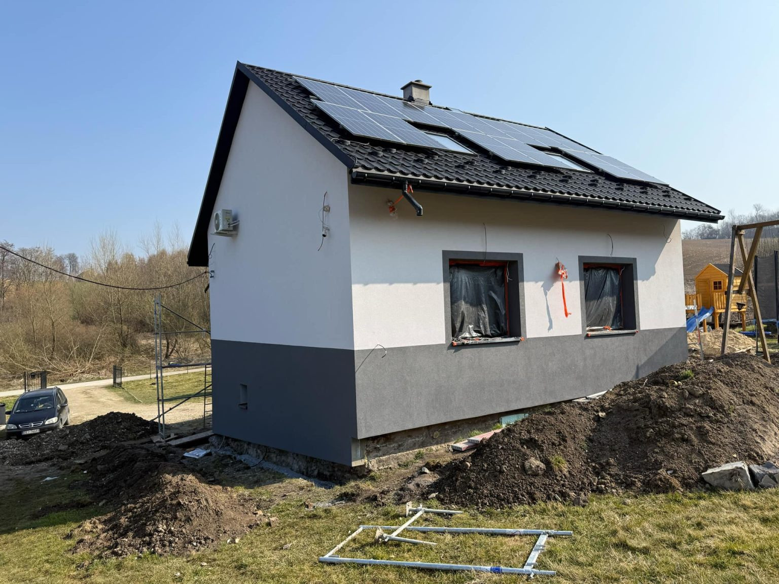 Dom po ociepleniu z panelami słonecznymi na dachu, okna zabezpieczone folią. Widoczne rusztowania i przygotowany teren wokół budynku. Dwukolorowa elewacja.