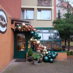 Handyman Tomasz Wawoczny - Dekoracja balonowa w odcieniach złota, brązu i zieleni zdobi wejście do cukierni Bonbon. Kompozycja tworzy łuk nad drzwiami, z roślinami w donicach u podstawy.