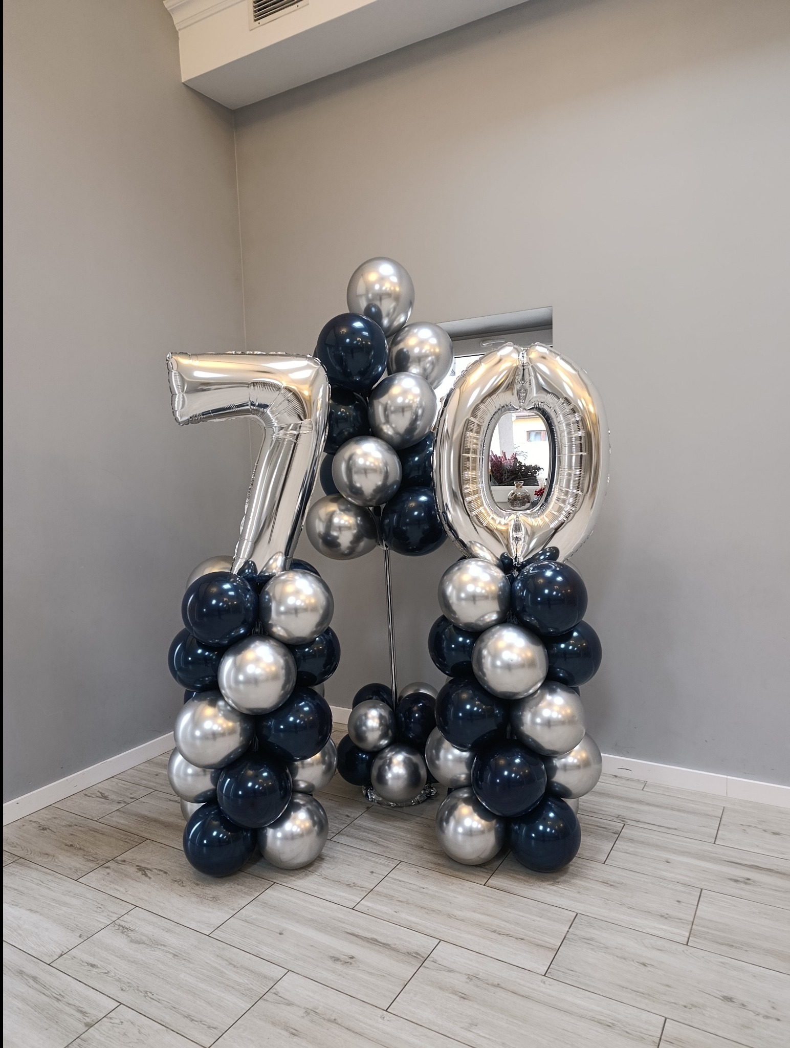 Dekoracja balonowa na 70. urodziny: srebrne cyfry z helem, czarne i srebrne balony ułożone w kolumny. Minimalistyczne tło, jasna podłoga.