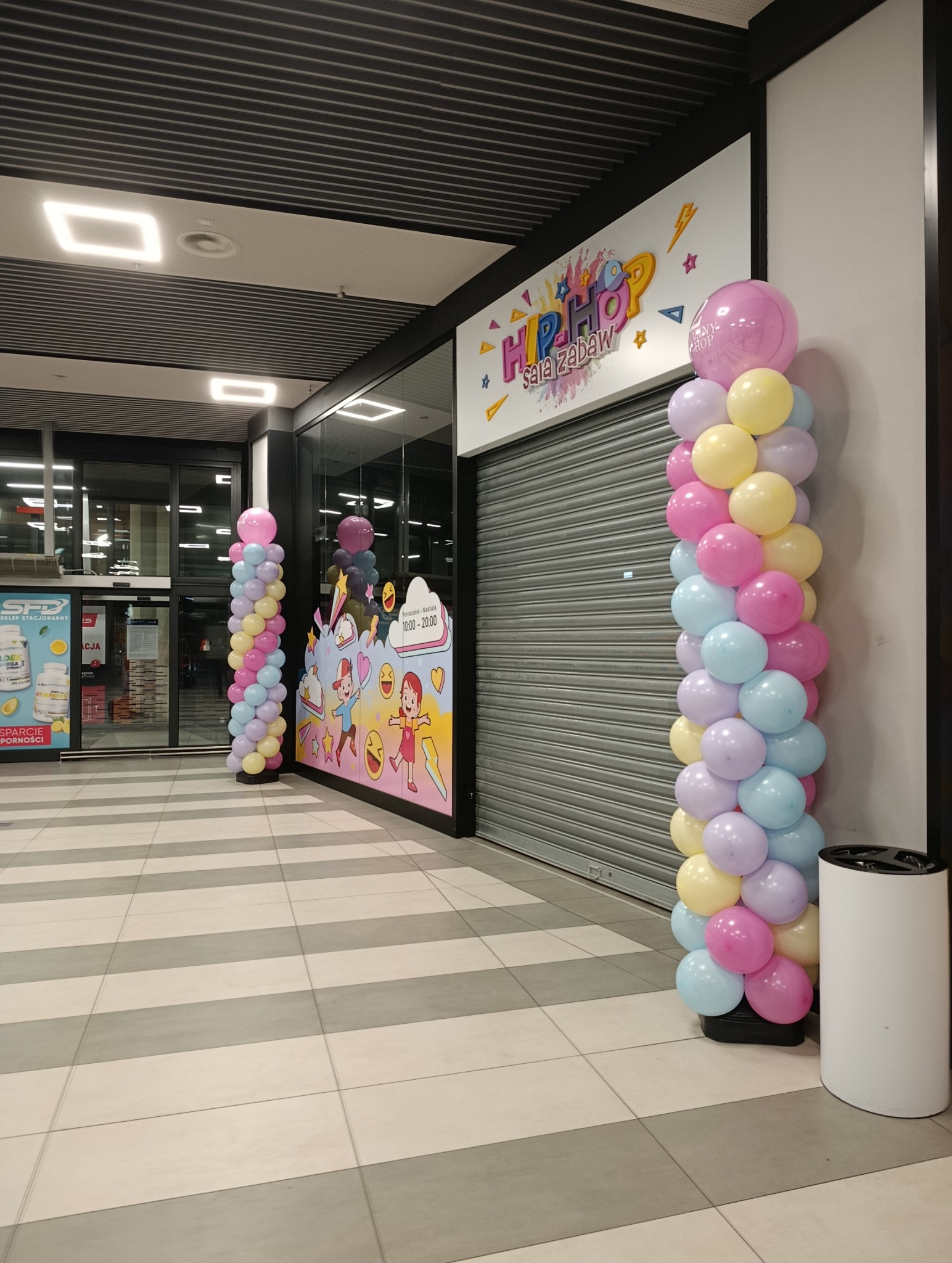 Kolorowe kolumny z balonów przy wejściu do sali zabaw 'Hip-Hop', z grafiką postaci z bajek na ścianie. Zamknięta roleta w tle. Dekoracja na imprezę.