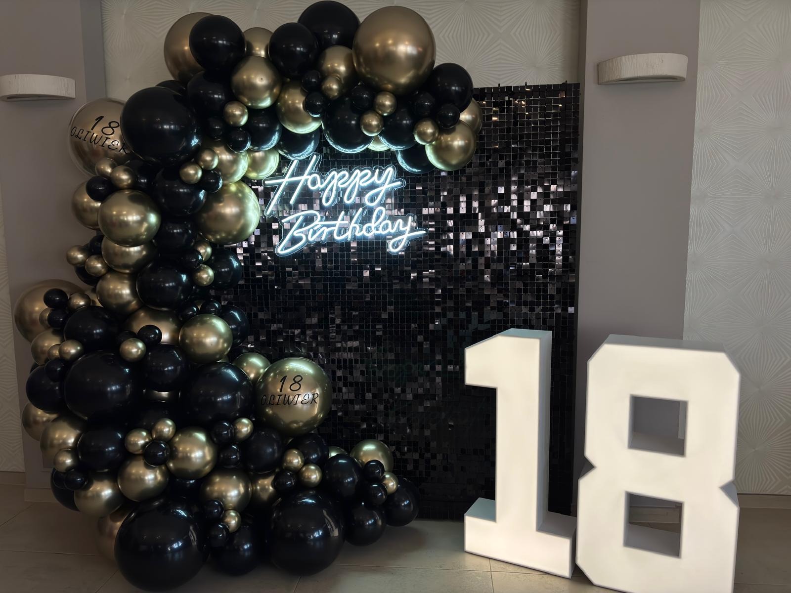 Dekoracja urodzinowa na osiemnastkę: czarno-złote balony, błyszcząca ścianka i neon 'Happy Birthday' tworzą elegancką oprawę imprezy.