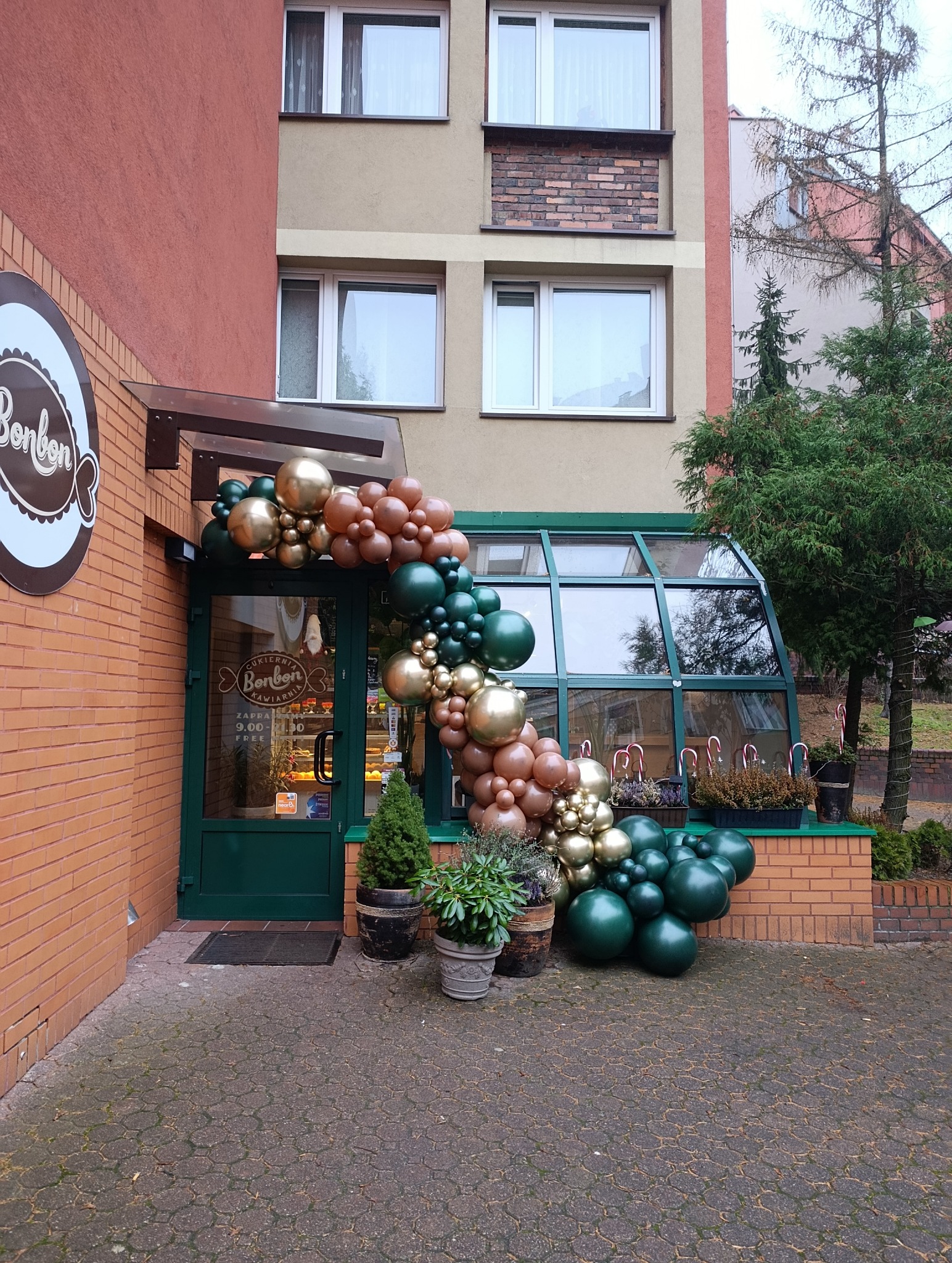 Dekoracja balonowa w odcieniach złota, brązu i zieleni zdobi wejście do cukierni Bonbon. Kompozycja tworzy łuk nad drzwiami, z roślinami w donicach u podstawy.