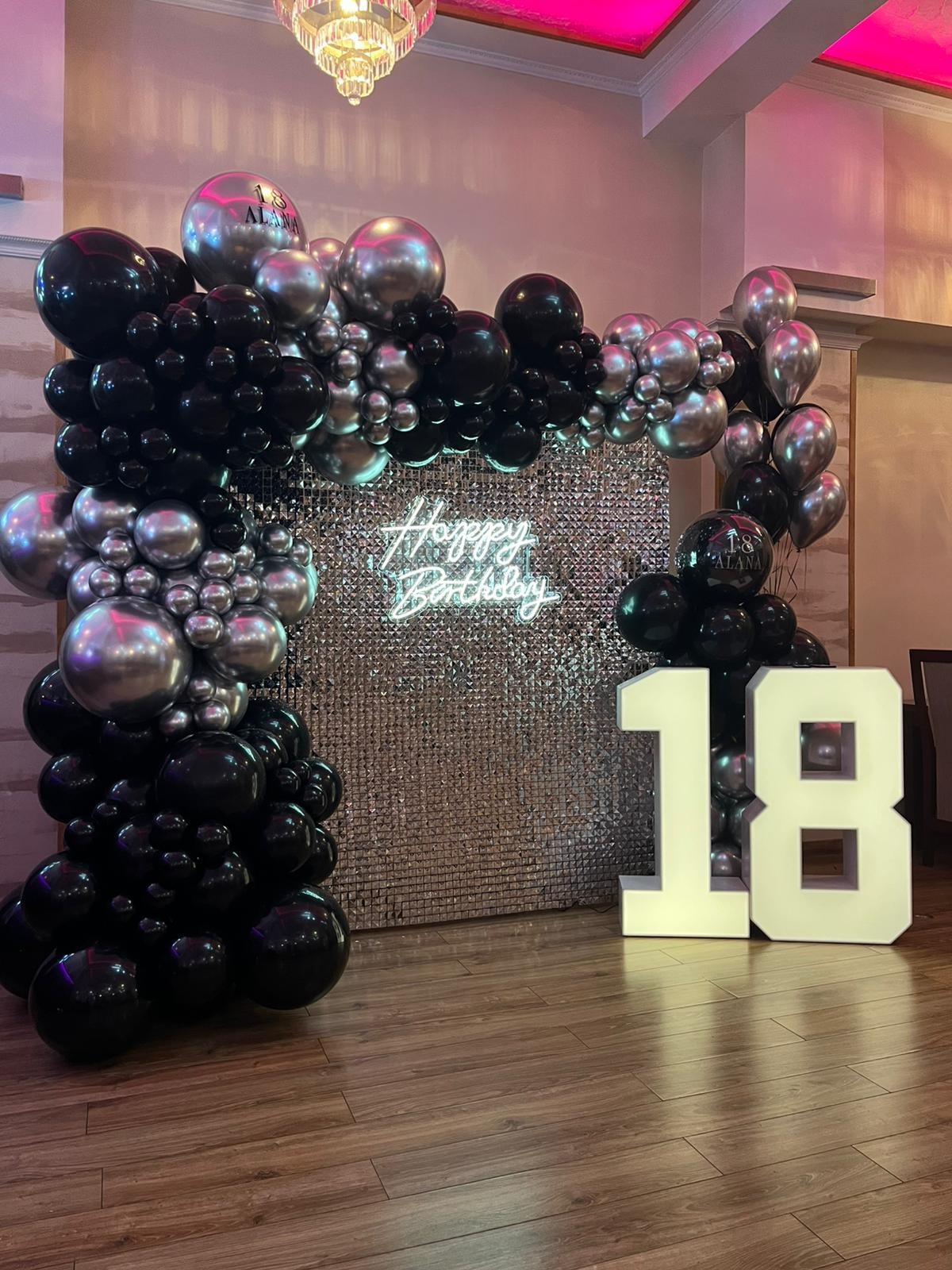 Dekoracja urodzinowa: srebrna ściana, neon 'Happy Birthday', czarno-srebrne balony i białe cyfry '18'. Elegancki wystrój na osiemnastkę.