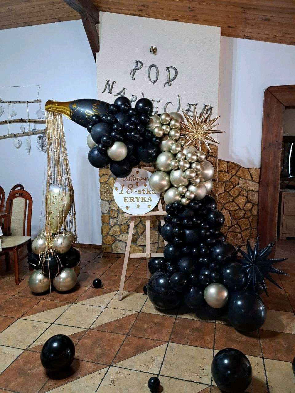 Dekoracja urodzinowa: butelka szampana z czarno-złotych balonów, gwiazdy i tabliczka z napisem '18-stka Eryka'.