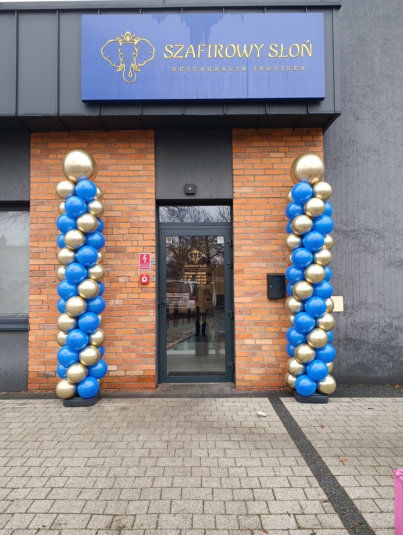 Dekoracja balonowa w kolorach niebieskim i złotym przed wejściem do restauracji indyjskiej 'Szafirowy Słoń'. Balony ułożone w kolumny po obu stronach drzwi.
