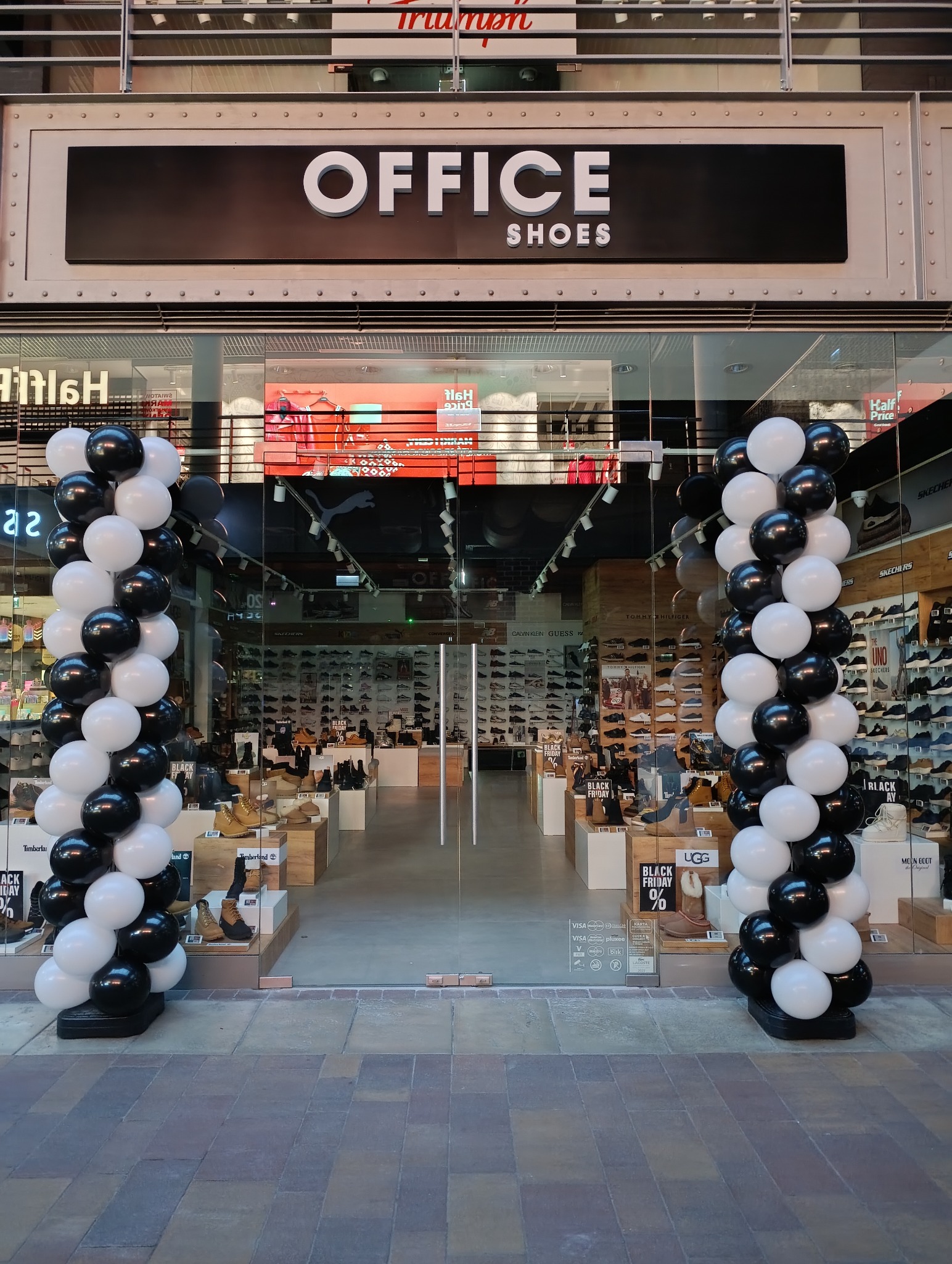 Wejście do sklepu obuwniczego 'Office Shoes' udekorowane kolumnami z czarno-białych balonów. W tle widoczne półki z obuwiem i plakaty promocyjne.