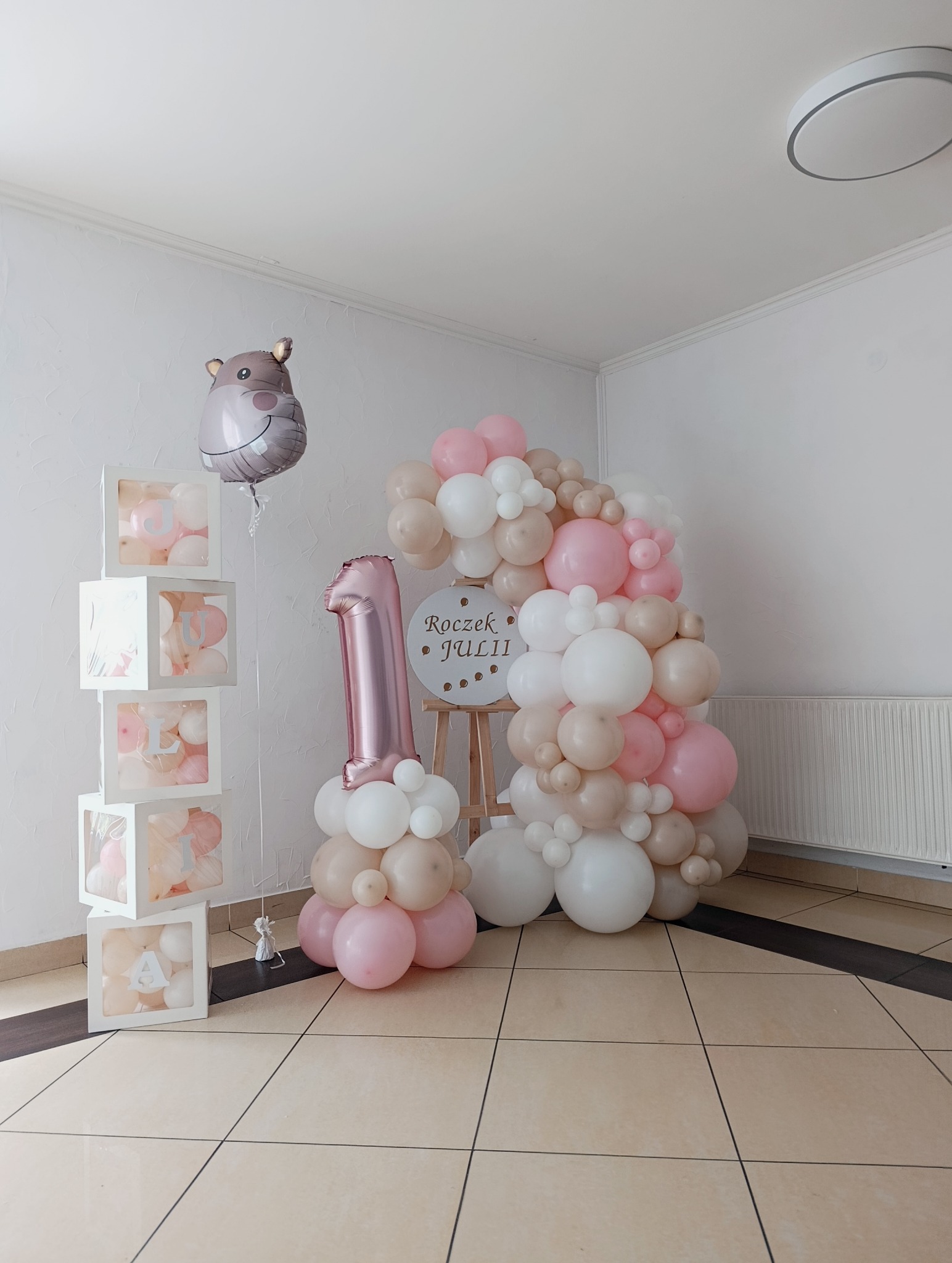 Dekoracja balonowa na roczek dziewczynki: różowe i białe balony, cyfra '1', balony w przezroczystych kostkach z literami imienia oraz balon w kształcie hipopotama.
