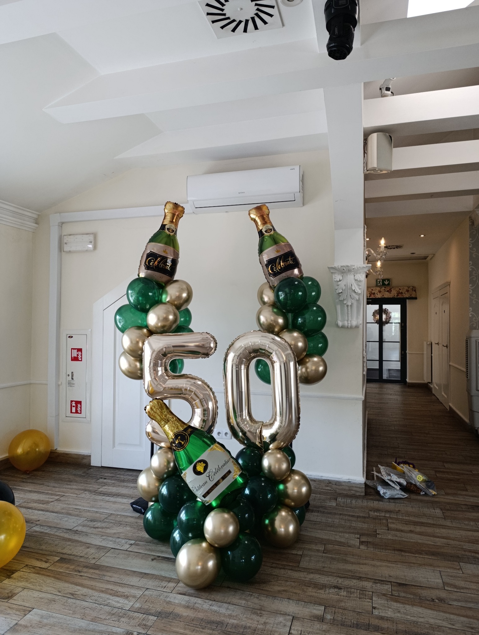 Dekoracja balonowa na 50 urodziny: balony w kształcie butelek szampana, cyfry '50' w kolorze złota i zielone balony. Dekoracja na drewnianej podłodze.