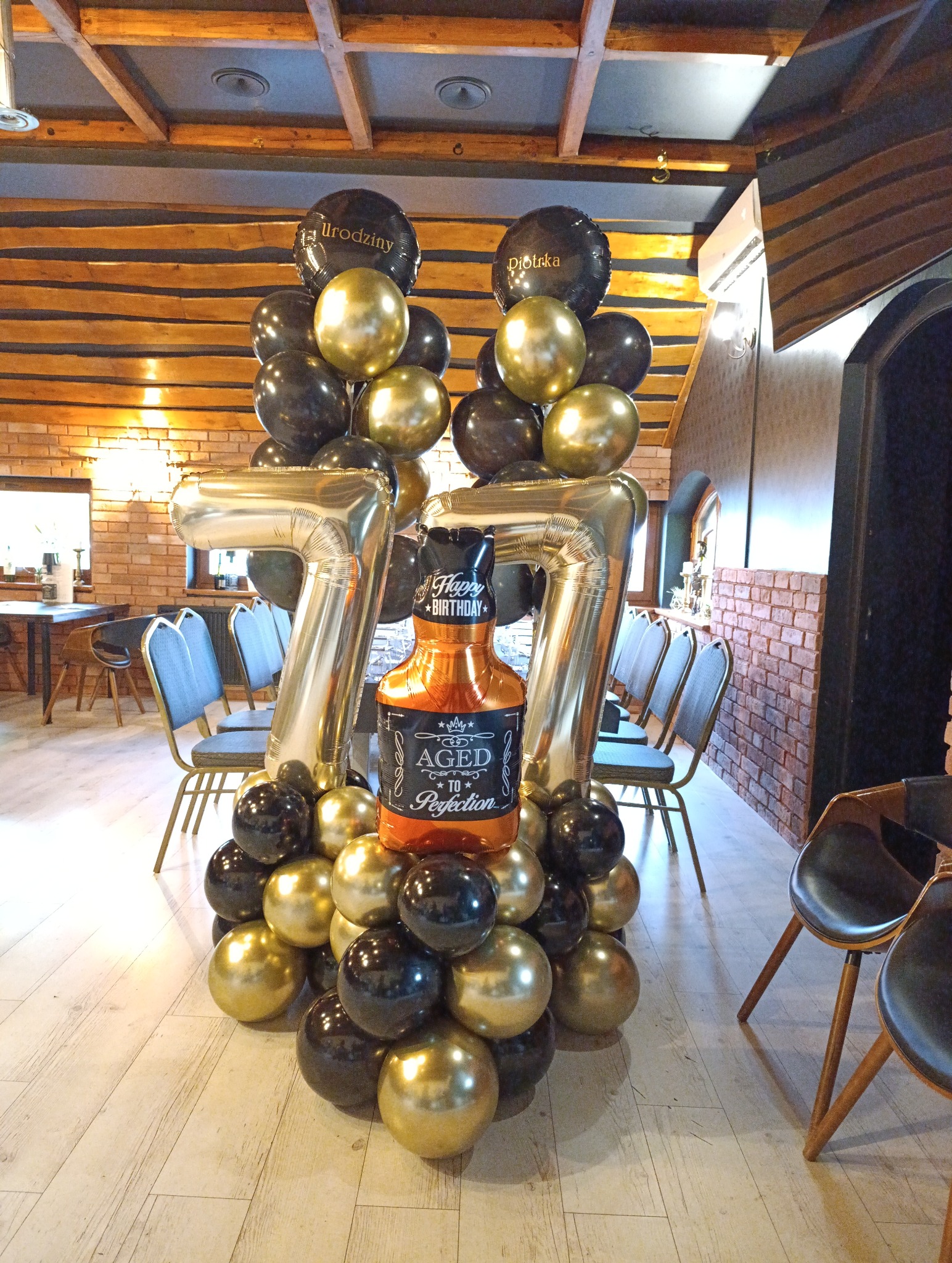 Dekoracja balonowa na 77 urodziny w restauracji: złote i czarne balony, cyfry '77' i balon w kształcie butelki whisky. Urodzinowe balony z napisem 'Urodziny' i 'Piotrka'.