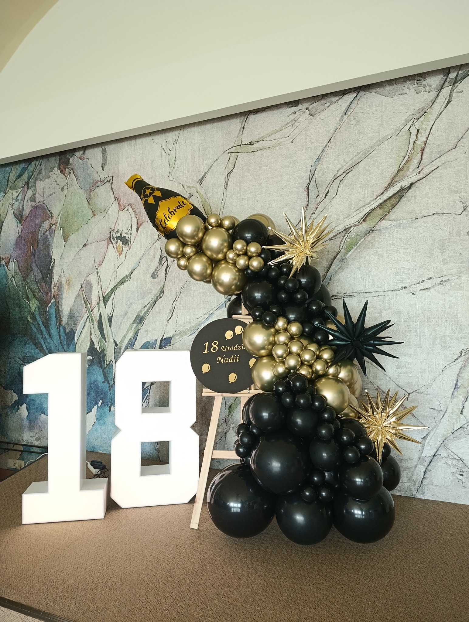 Dekoracja balonowa na 18 urodziny Nadii: czarne i złote balony, butelka szampana, gwiazdy, tabliczka z imieniem i wiekiem, białe cyfry '18'.