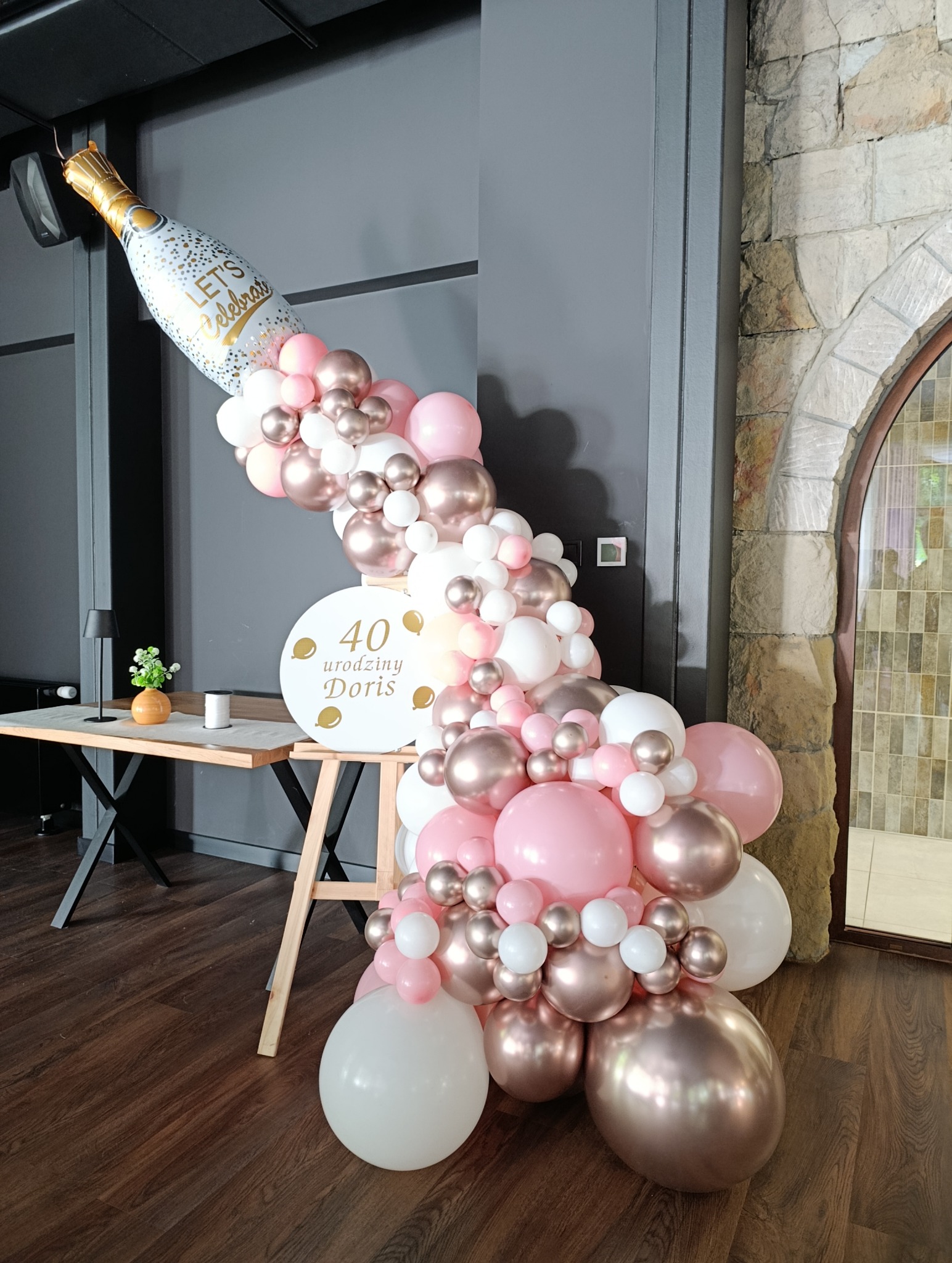 Dekoracja balonowa na 40. urodziny Doris: butelka szampana z balonów w odcieniach różu, bieli i złota, ustawiona obok stołu z kwiatami i świecami.