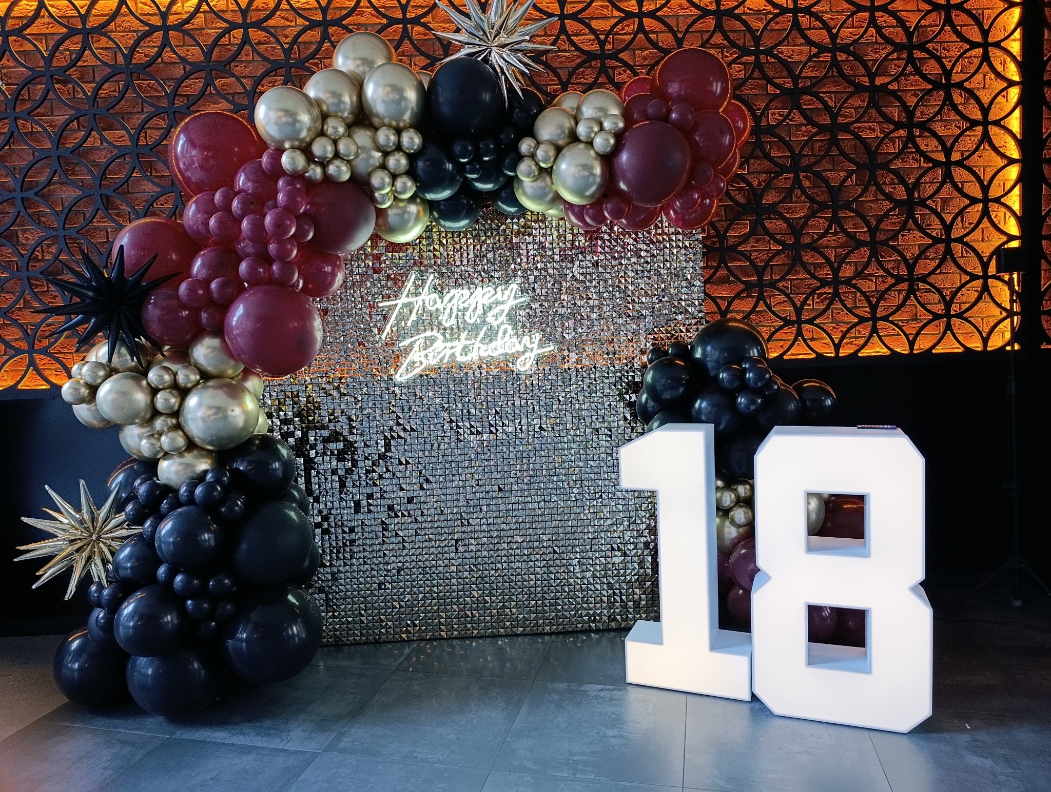 Dekoracja urodzinowa: balony w odcieniach bordo, czerni i złota tworzą łuk wokół błyszczącej ścianki z napisem neonowym 'Happy Birthday' i cyfrą 18.