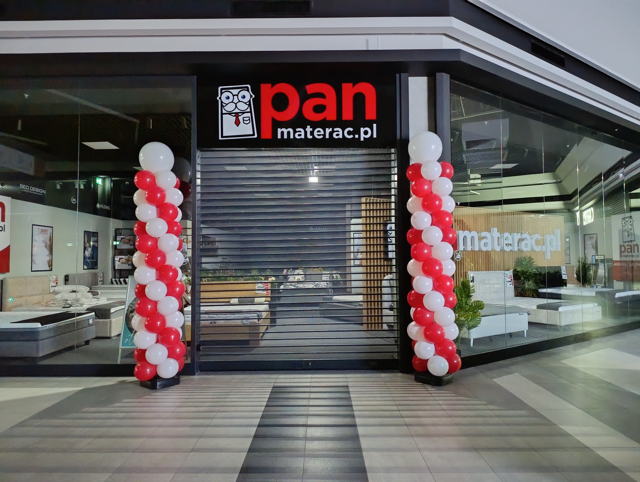Otwarcie sklepu z materacami: kolumny z czerwono-białych balonów flankują wejście z logiem 'panmaterac.pl'. W tle ekspozycja materacy za zamkniętą roletą.