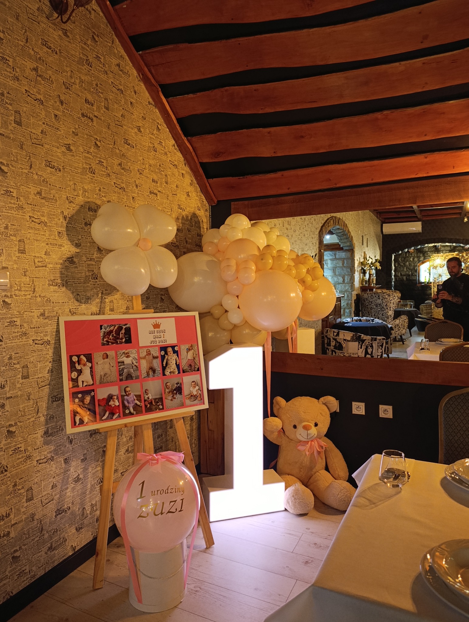 Dekoracja na roczek: balon z napisem '1 urodziny Zuzi', podświetlana cyfra '1', miś, tablica ze zdjęciami dziecka, kompozycja z białych i beżowych balonów.