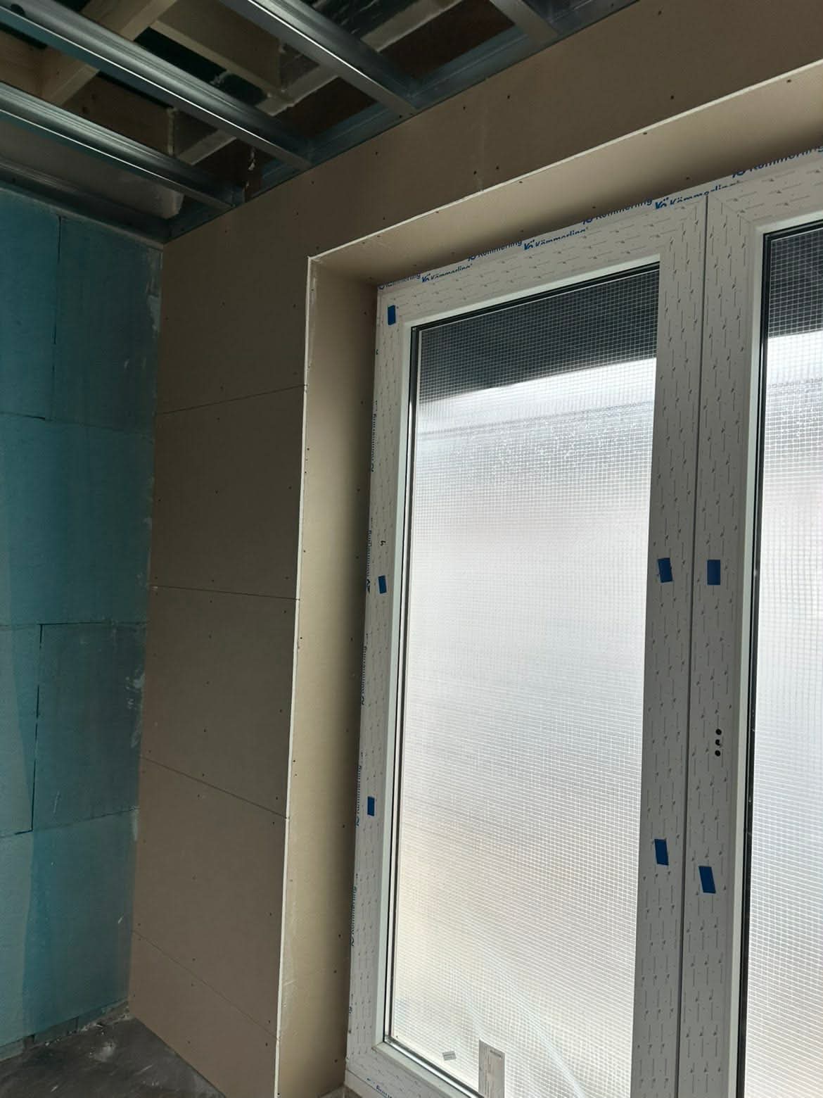 Montaż płyty G-K wokół okna w surowym wnętrzu. Widoczne profile sufitowe i ściana z bloczków. Etap prac wykończeniowych, przygotowanie do dalszych prac.