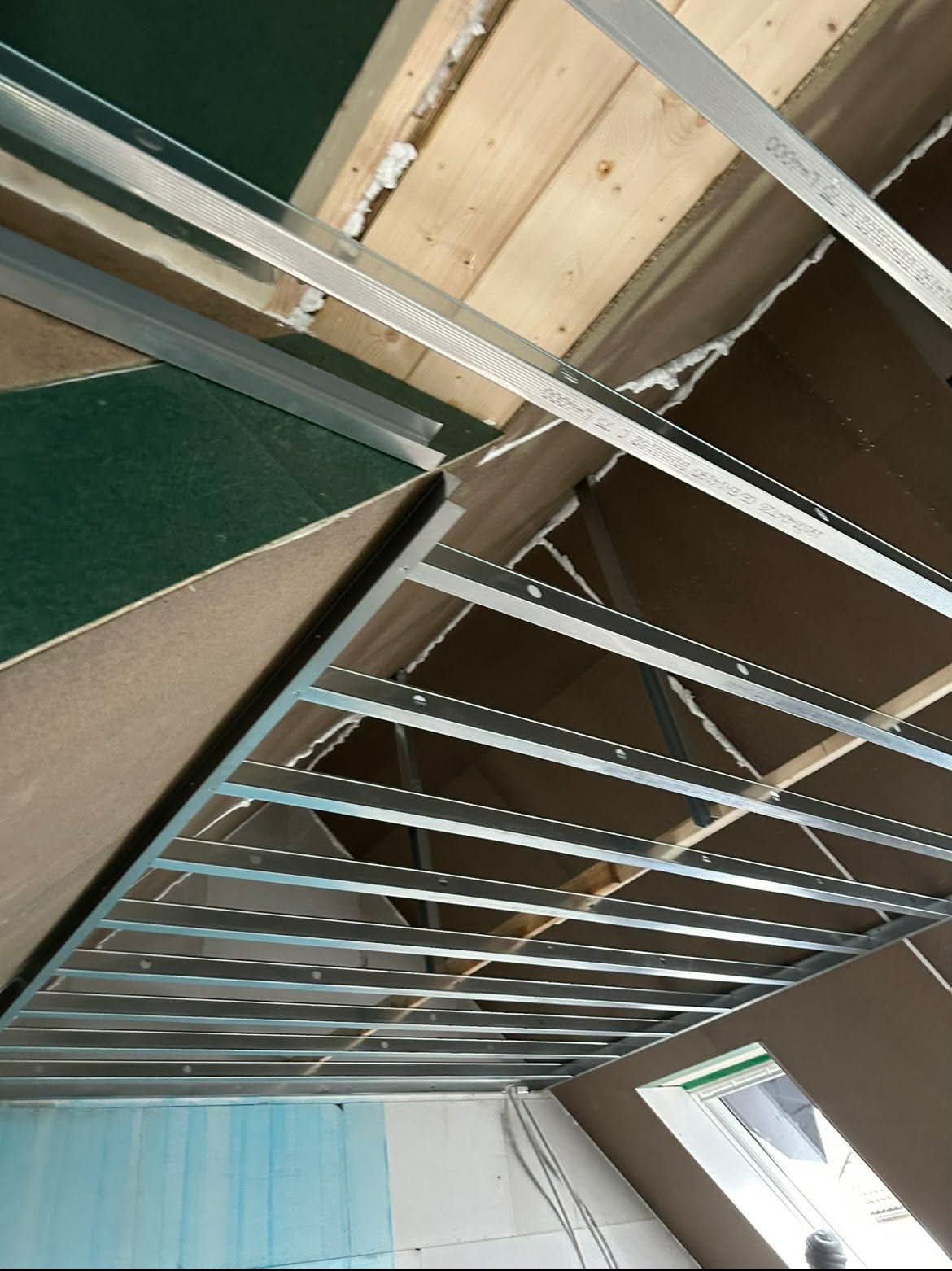 Sufit podwieszany z metalową konstrukcją w trakcie montażu, widoczne płyty karton-gips i drewniane elementy konstrukcyjne poddasza. Ujęcie z dołu, w tle okno.