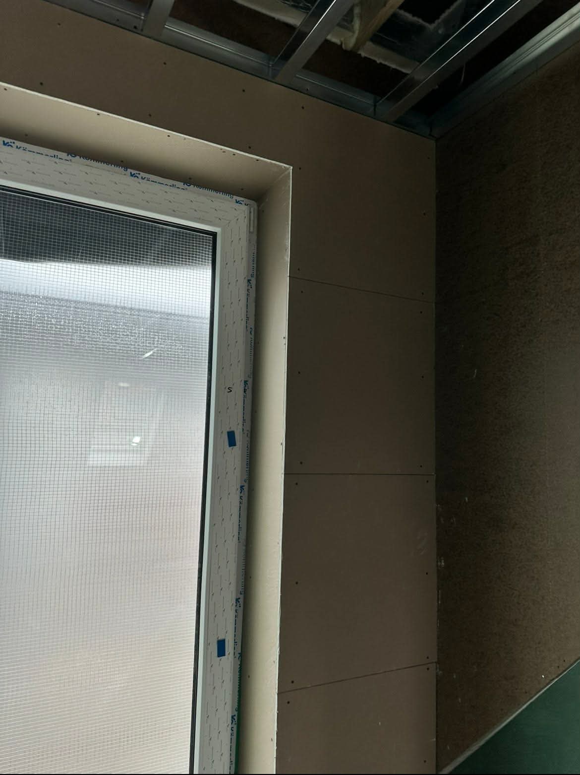 Montaż płyty gipsowo-kartonowej przy oknie, widoczne profile metalowe sufitu. Ściana z płyt, okno z folią ochronną. Etap prac wykończeniowych.
