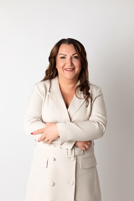 Uśmiechnięta kobieta w beżowej sukience-marynarce, z rękami skrzyżowanymi na wysokości talii, na jasnym tle. Portret doradcy w neutralnym otoczeniu.