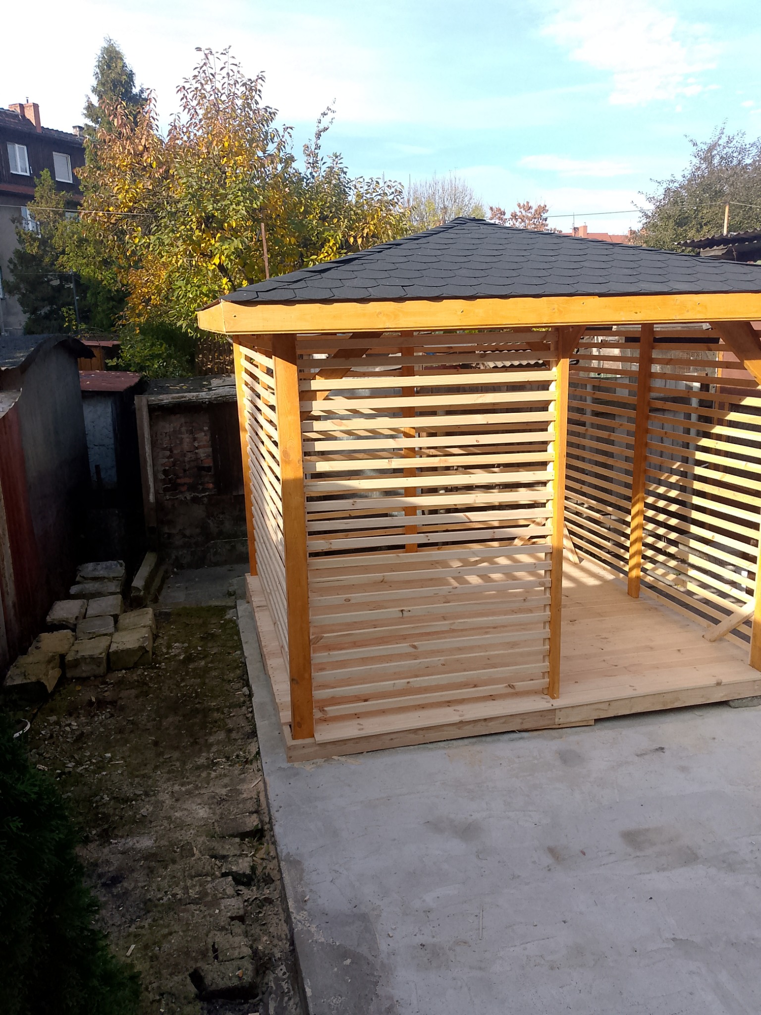 Nowa, jasna altana z drewnianymi lamelami i czarnym dachem, widziana z góry na betonowym podłożu. W tle drzewa i budynki.