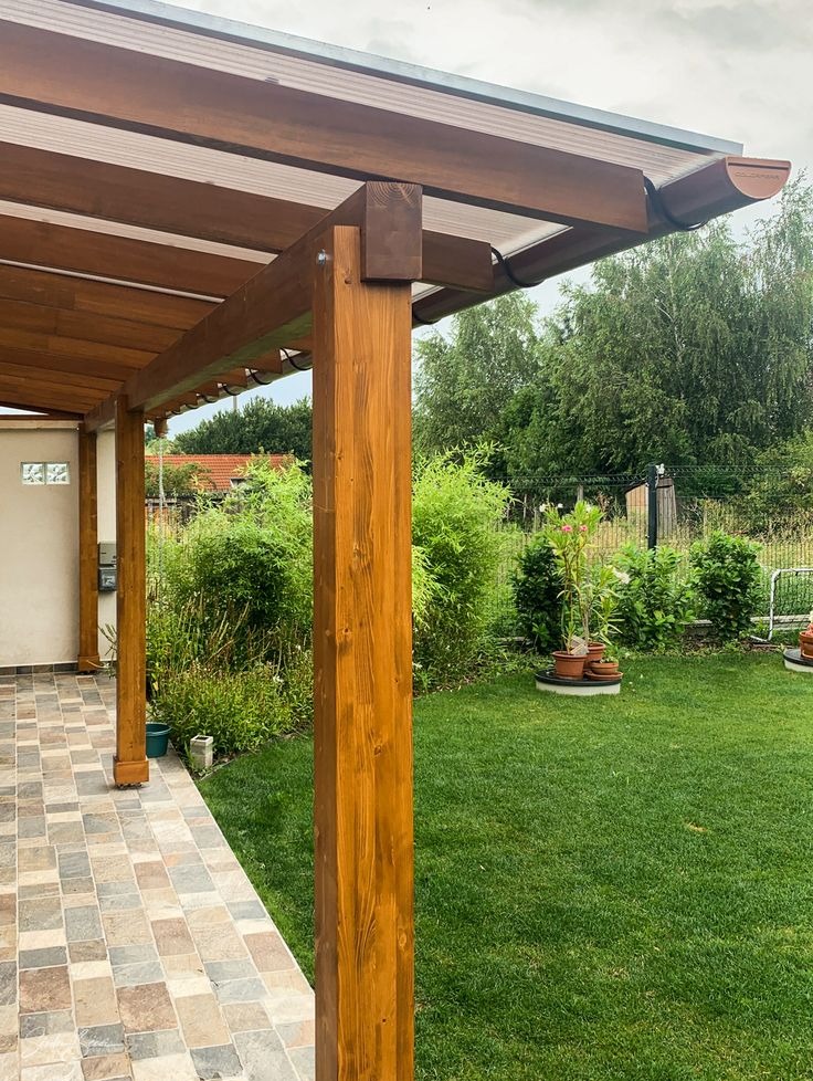 Drewniana pergola z brązowymi słupami i przezroczystym zadaszeniem, widok z tarasu na zielony ogród. Detal konstrukcji i otoczenia.