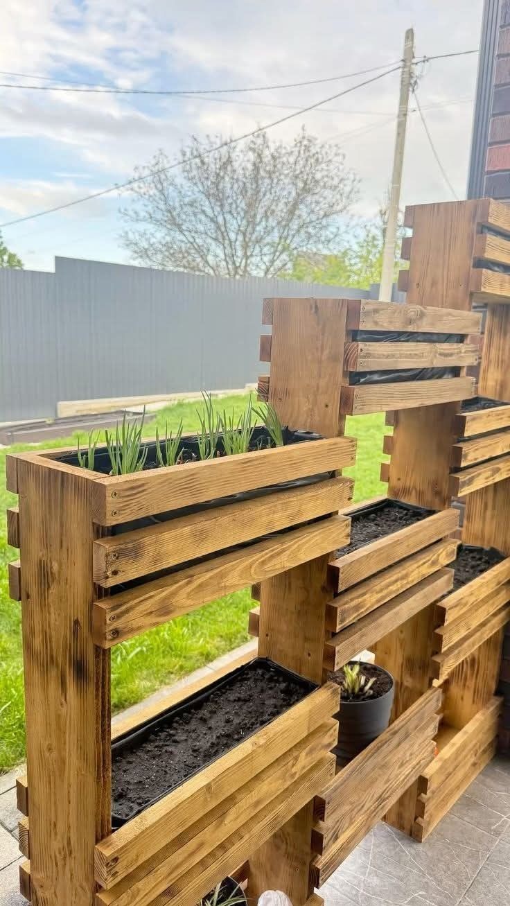 Drewniana konstrukcja z donicami, tworząca pionowy ogród. Widoczne rośliny w jednej z donic, tło z szarym ogrodzeniem i zielenią. Ciepłe oświetlenie podkreśla strukturę drewna.