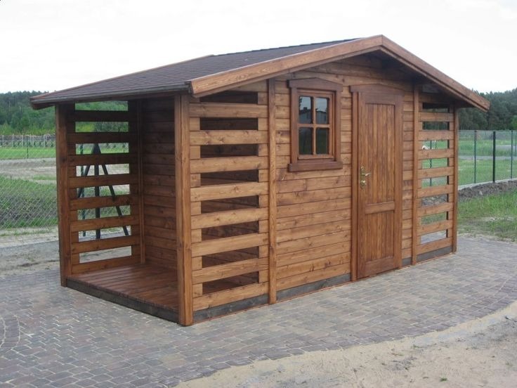 Drewniana konstrukcja z zadaszeniem, z oknem i drzwiami, otoczona ażurowymi ścianami. Widoczna faktura drewna i brązowy kolor impregnatu. Całość na tle zieleni.