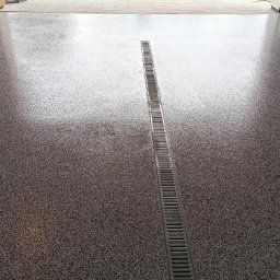 Bastix - Błyszcząca, szara posadzka żywiczna z odpływem liniowym na środku, wykonana w garażu. Widoczne zabezpieczenia taśmą malarską przy ścianach.