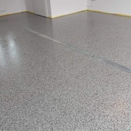 Bastix - Błyszcząca, szara posadzka żywiczna z odpływem liniowym w nowym pomieszczeniu. Ściany zabezpieczone taśmą malarską. Minimalistyczny design.