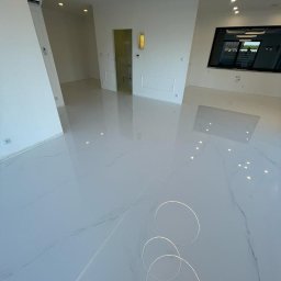 Bastix - Błyszcząca, biała posadzka z efektem marmuru i dekoracyjnymi okręgami w przestronnym, minimalistycznym wnętrzu z oknem i oświetleniem sufitowym.