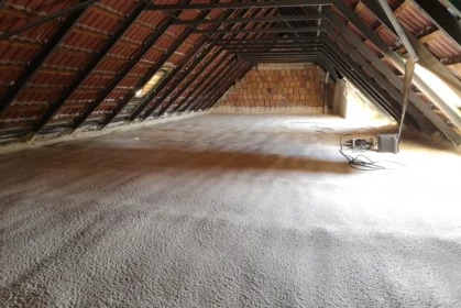 Poddasze z widoczną warstwą izolacji natryskowej, drewniana konstrukcja dachu i fragment murowanej ściany w tle. Widoczne okno dachowe i sprzęt do natrysku.