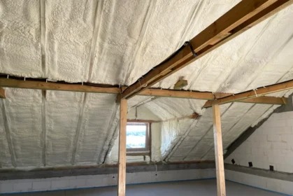 Poddasze z drewnianą konstrukcją pokryte grubą warstwą jasnej pianki izolacyjnej, widoczne okno i fragment ściany z pustaków. Realizacja ocieplenia.