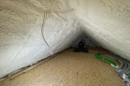 Poddasze ocieplone pianką PUR, widoczne przewody i rury. Biała, porowata struktura pianki pokrywa skośne ściany i sufit. Podłoga z płyt OSB.