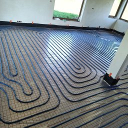 KaBi Hydraulika - Niebieskie rury ogrzewania podłogowego ułożone na siatce w jasnym pomieszczeniu z oknami. Widoczne czarne taśmy mocujące rury do folii. Biały filar w prawym dolnym rogu.