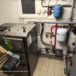 KaBi Hydraulika KAROL BILSKI - Nowoczesny piec CO marki Kolton z podajnikiem, połączony z instalacją grzewczą. Widoczne rury, zawory i zbiorniki wyrównawcze. Całość w kotłowni z wyłożoną terakotą.