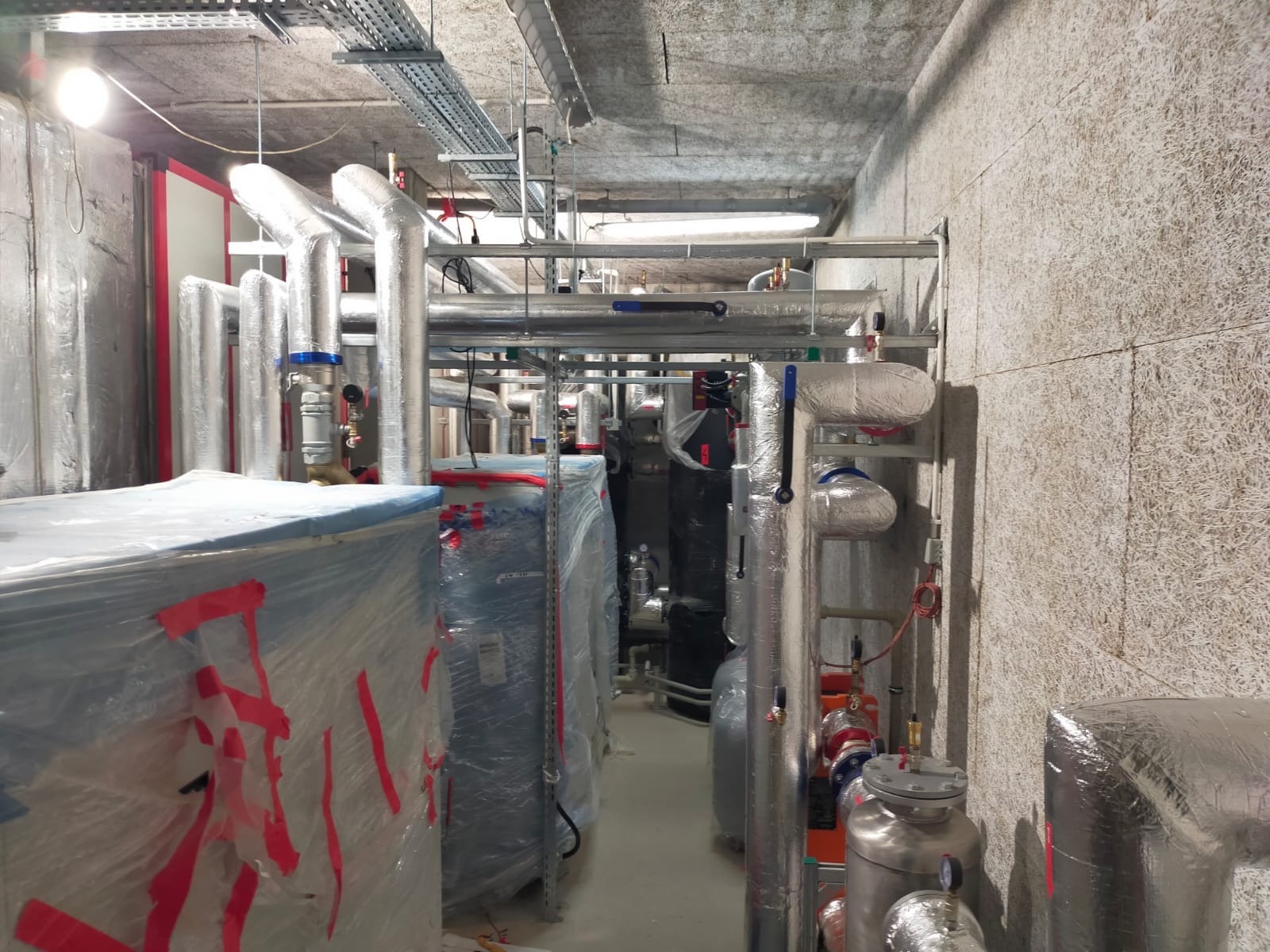 Węzeł cieplny w piwnicy: rury w izolacji, zawory, zbiorniki, instalacje pod stropem. Elementy zabezpieczone folią. Widok na instalację w trakcie prac.