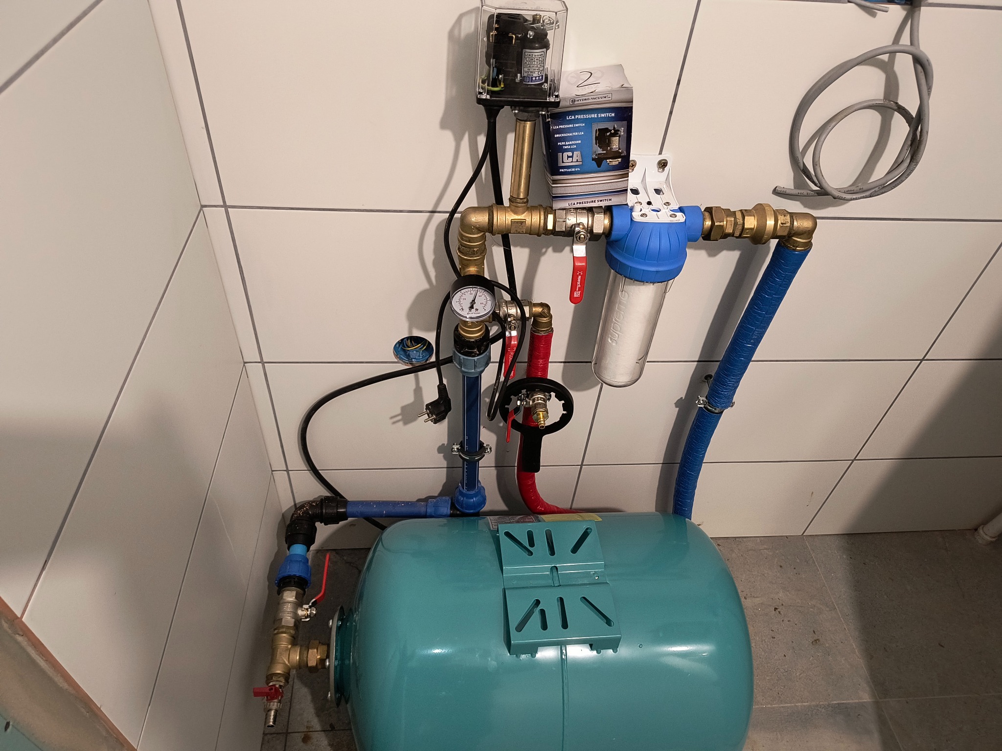 Niebieski hydrofor z osprzętem (manometr, zawory, filtr) na tle białych płytek. Widoczna skrzynka sterownicza i opakowanie LCA Pressure Switch.