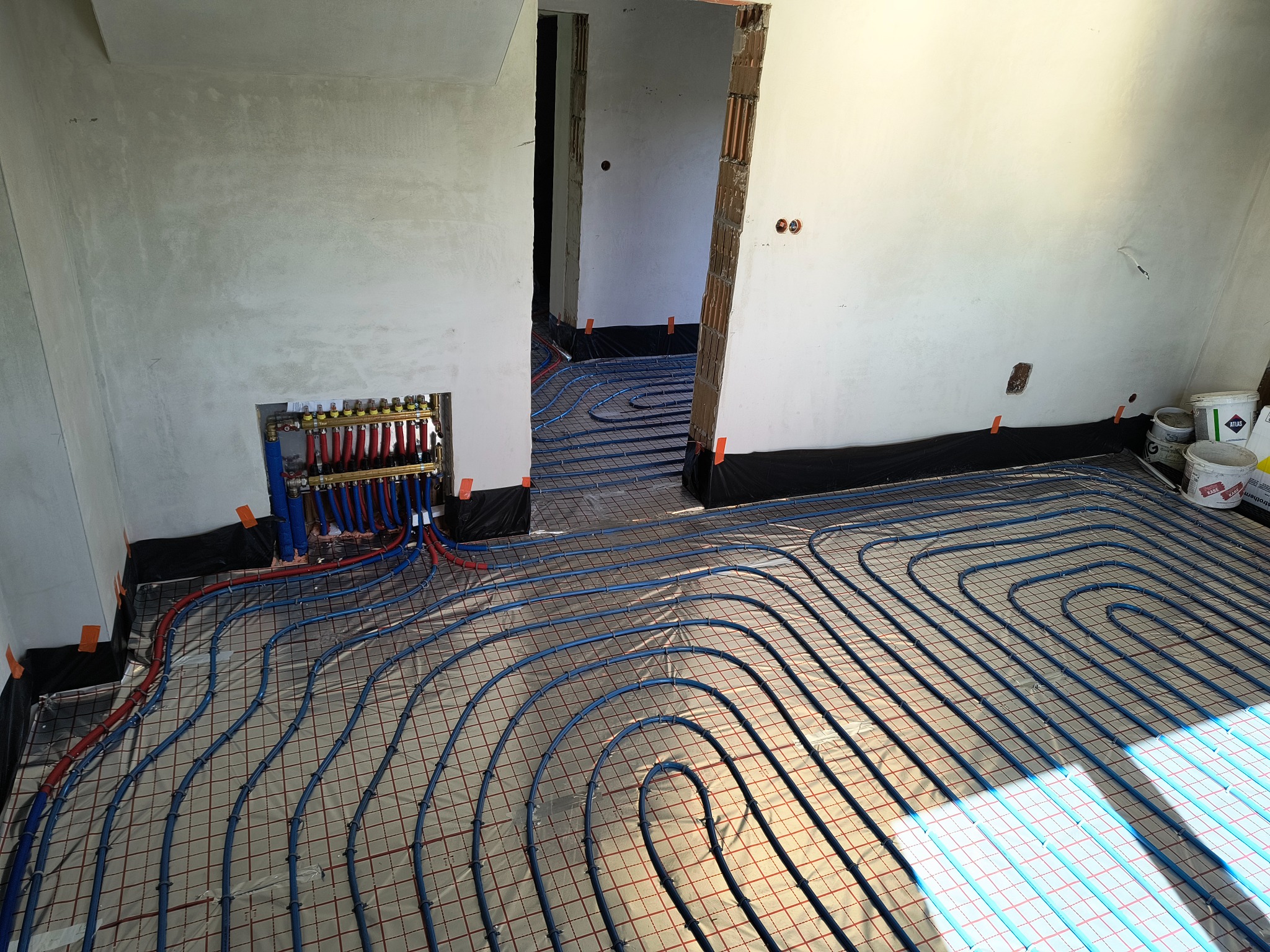 Niebieskie rury ogrzewania podłogowego ułożone na folii z siatką, widoczne kolektory i fragmenty ścian w surowym stanie. Instalacja przed wylaniem posadzki.
