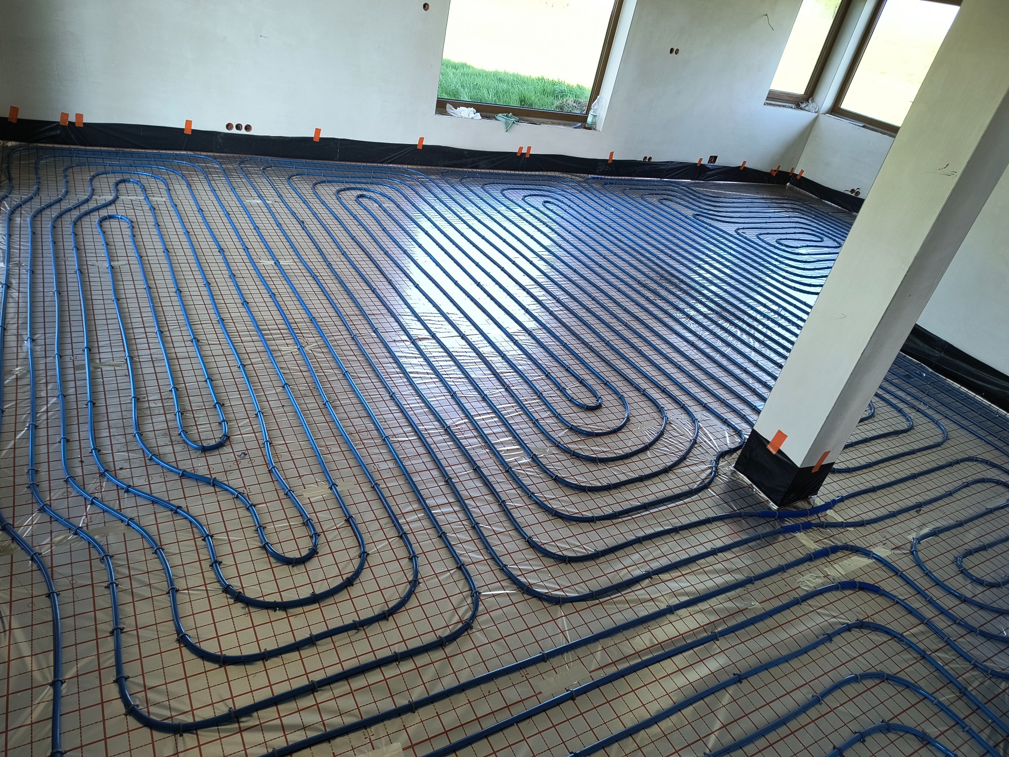 Niebieskie rury ogrzewania podłogowego ułożone na siatce w jasnym pomieszczeniu z oknami. Widoczne czarne taśmy mocujące rury do folii. Biały filar w prawym dolnym rogu.