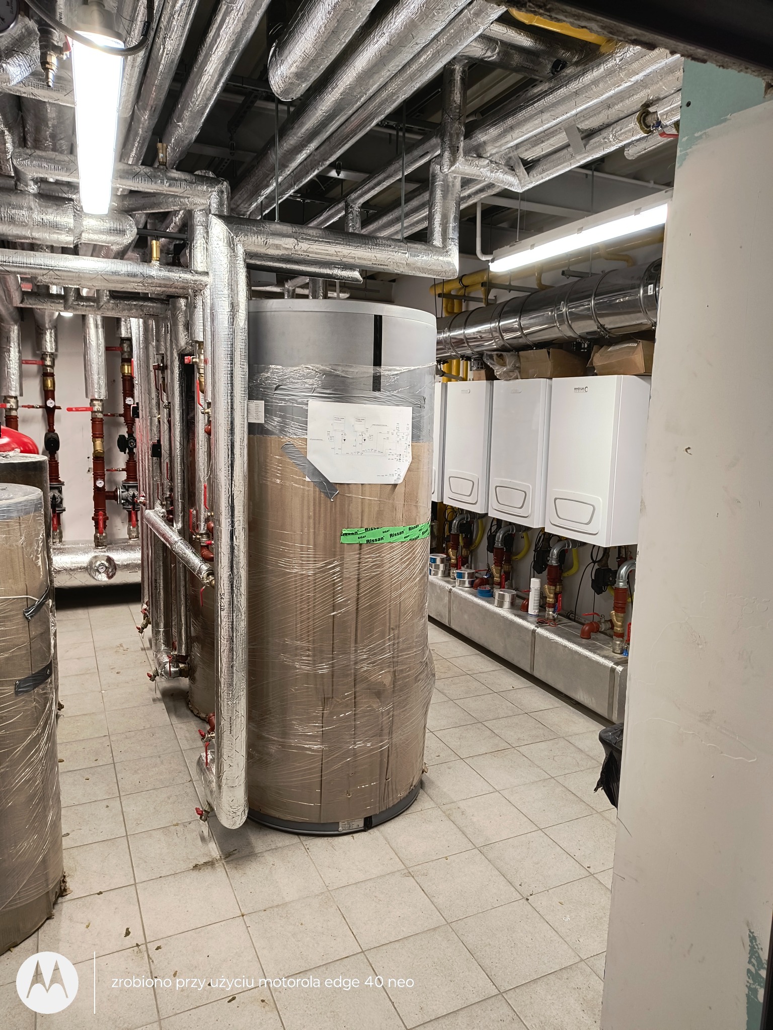 Kotłownia z nowymi piecami gazowymi i zbiornikami, rury w izolacji termicznej. Widok na urządzenia przed uruchomieniem, w folii ochronnej.