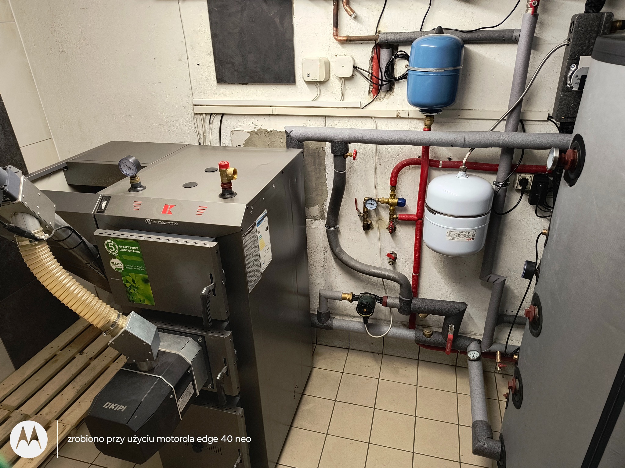 Nowoczesny piec CO marki Kolton z podajnikiem, połączony z instalacją grzewczą. Widoczne rury, zawory i zbiorniki wyrównawcze. Całość w kotłowni z wyłożoną terakotą.