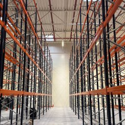 VAN BAU - Puste, pomarańczowo-czarne regały magazynowe w hali, widok z perspektywy podłogi, symetryczny układ, oświetlenie sufitowe.