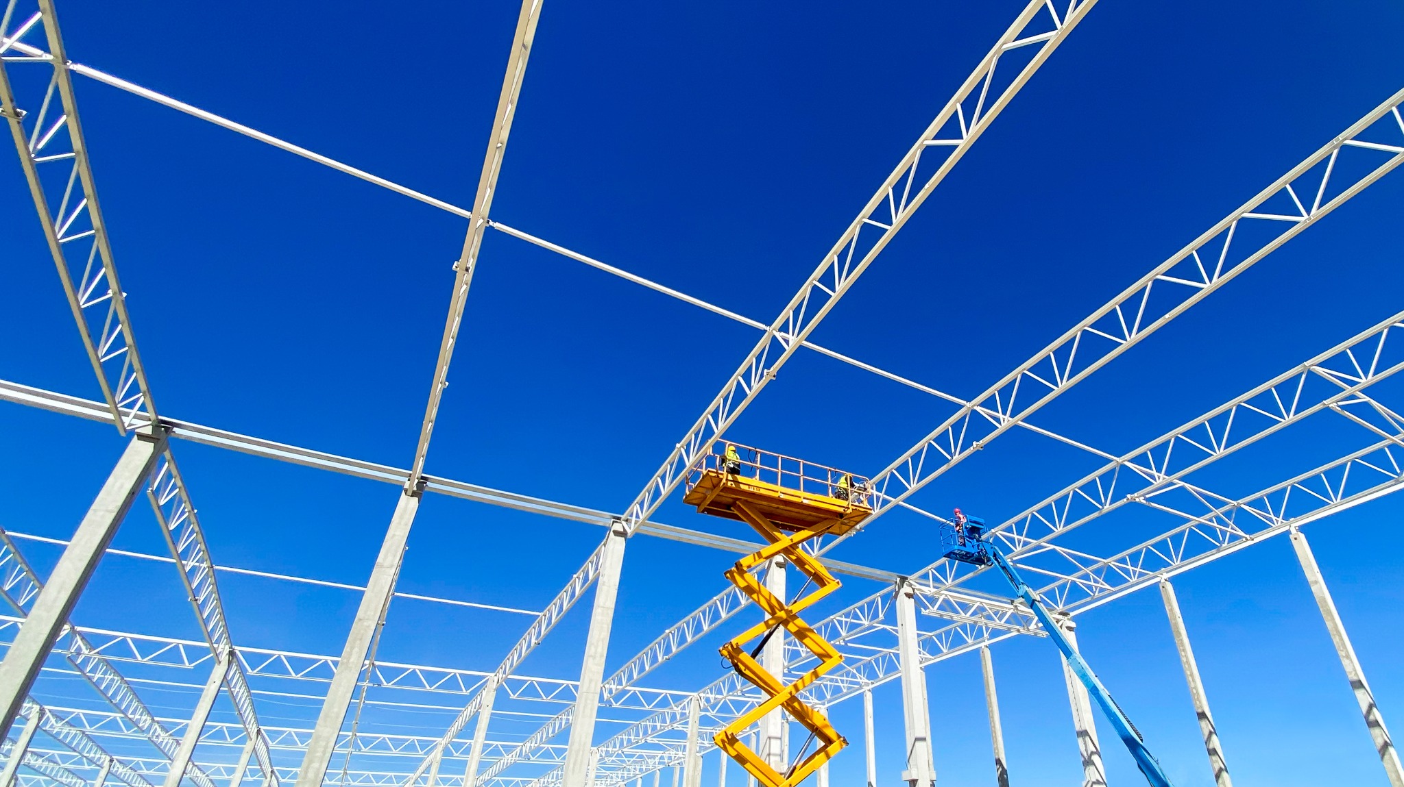 Montaż konstrukcji stalowej hali z użyciem podnośników koszowych na tle błękitnego nieba. Widok z dołu, podkreślający rozmach budowy.