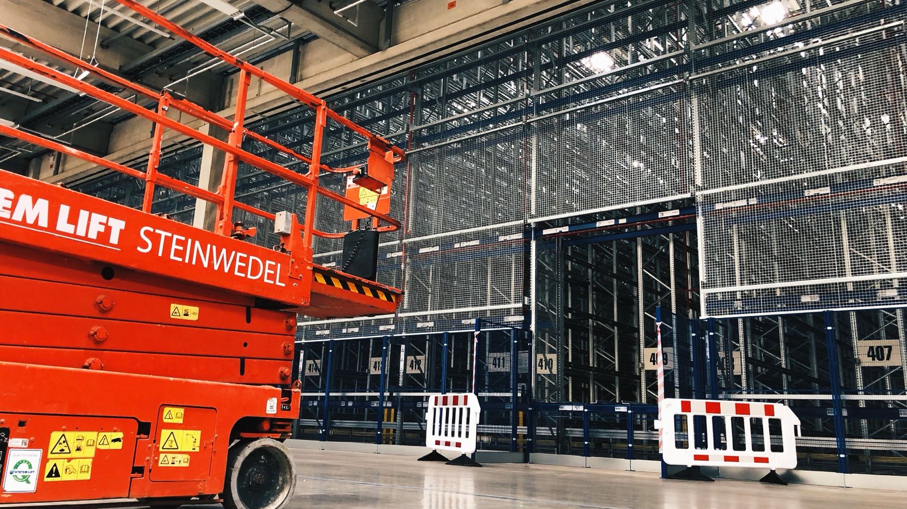 Częściowy widok na wysokie regały magazynowe z siatki, z pomarańczowym podnośnikiem platformowym po lewej i barierkami ochronnymi na pierwszym planie.