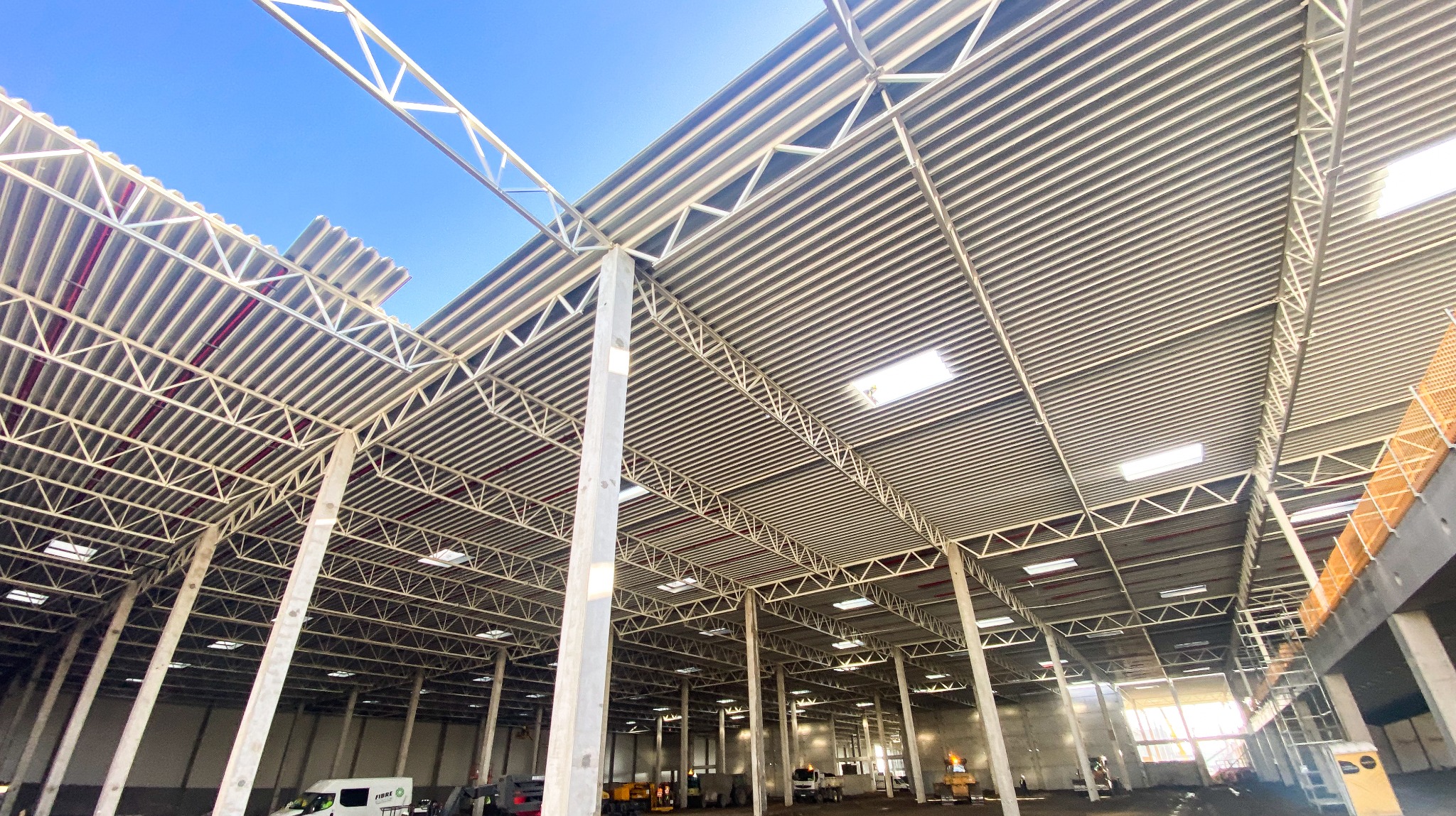 Wnętrze hali w budowie z widoczną konstrukcją stalową dachu i słupami nośnymi. Częściowo zamontowane płyty dachowe, w tle sprzęt budowlany i pojazdy.