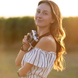 Anastazja Miketa Digital Branding - Uśmiechnięta brunetka w sukience w paski, z aparatem retro na szyi, pozuje w plenerze w ciepłym, zachodzącym słońcu. Delikatne światło podkreśla jej włosy.