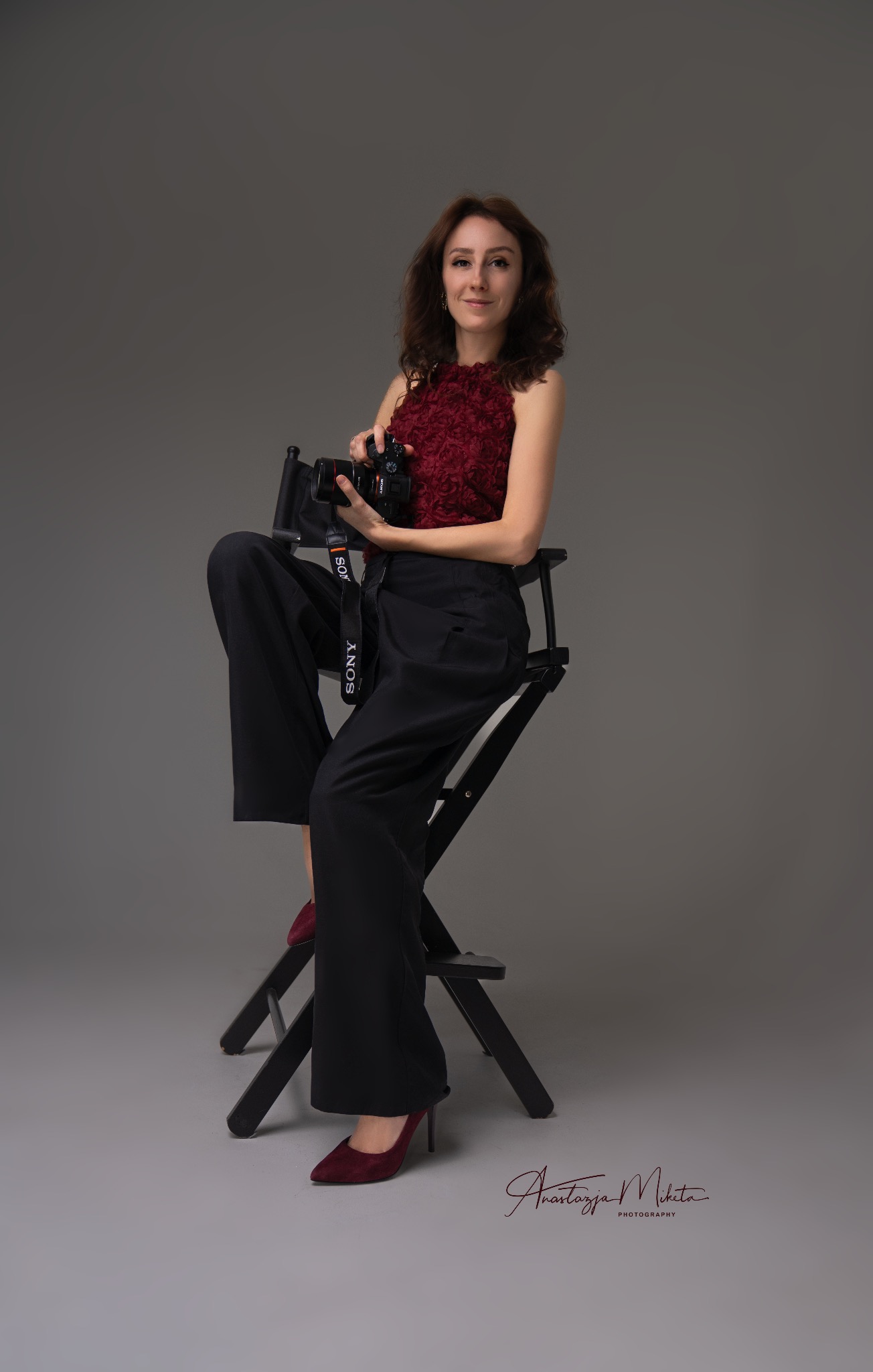 Kobieta z aparatem Sony siedzi na czarnym krześle reżyserskim na szarym tle. Ubrana w bordową bluzkę i czarne spodnie. Czerwone szpilki. Sygnatura fotografa w prawym dolnym rogu.