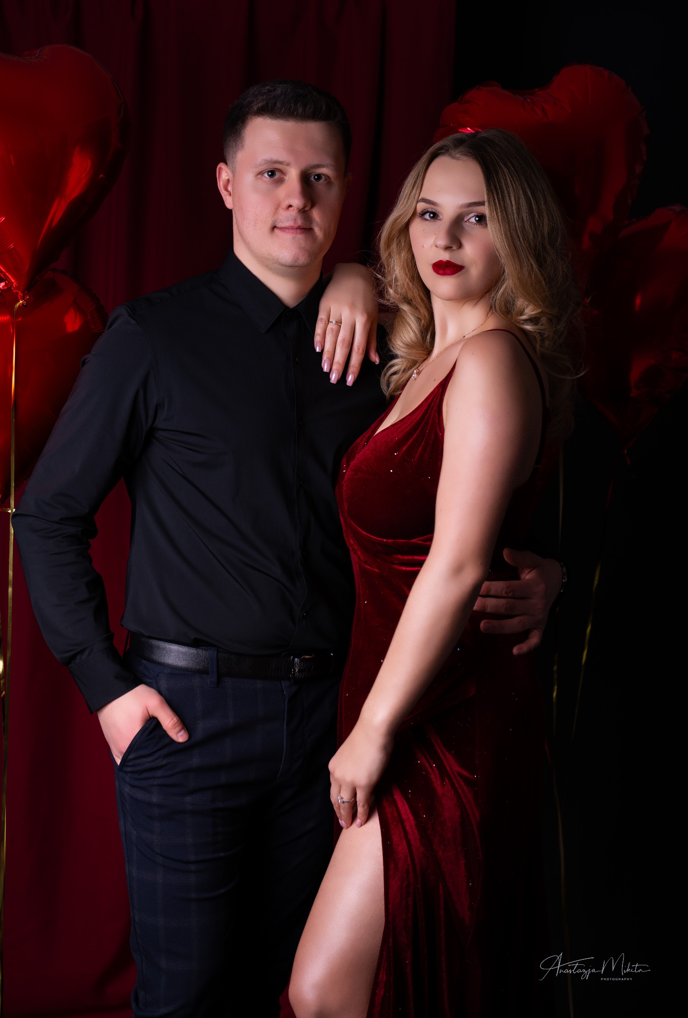 Elegancka sesja narzeczeńska w Żorach: para pozuje na tle czerwonych balonów w kształcie serc. Kobieta w aksamitnej sukni z rozcięciem, mężczyzna w czarnej koszuli.