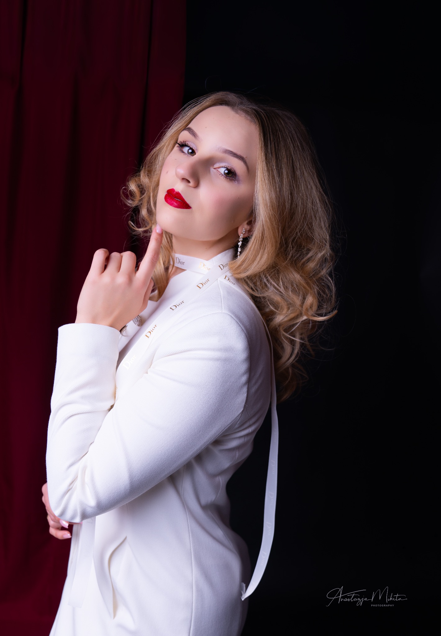 Portret kobiety w białym żakiecie Dior na tle czerwonej zasłony i czerni. Usta pomalowane czerwoną szminką, włosy blond, kręcone. Delikatna biżuteria.