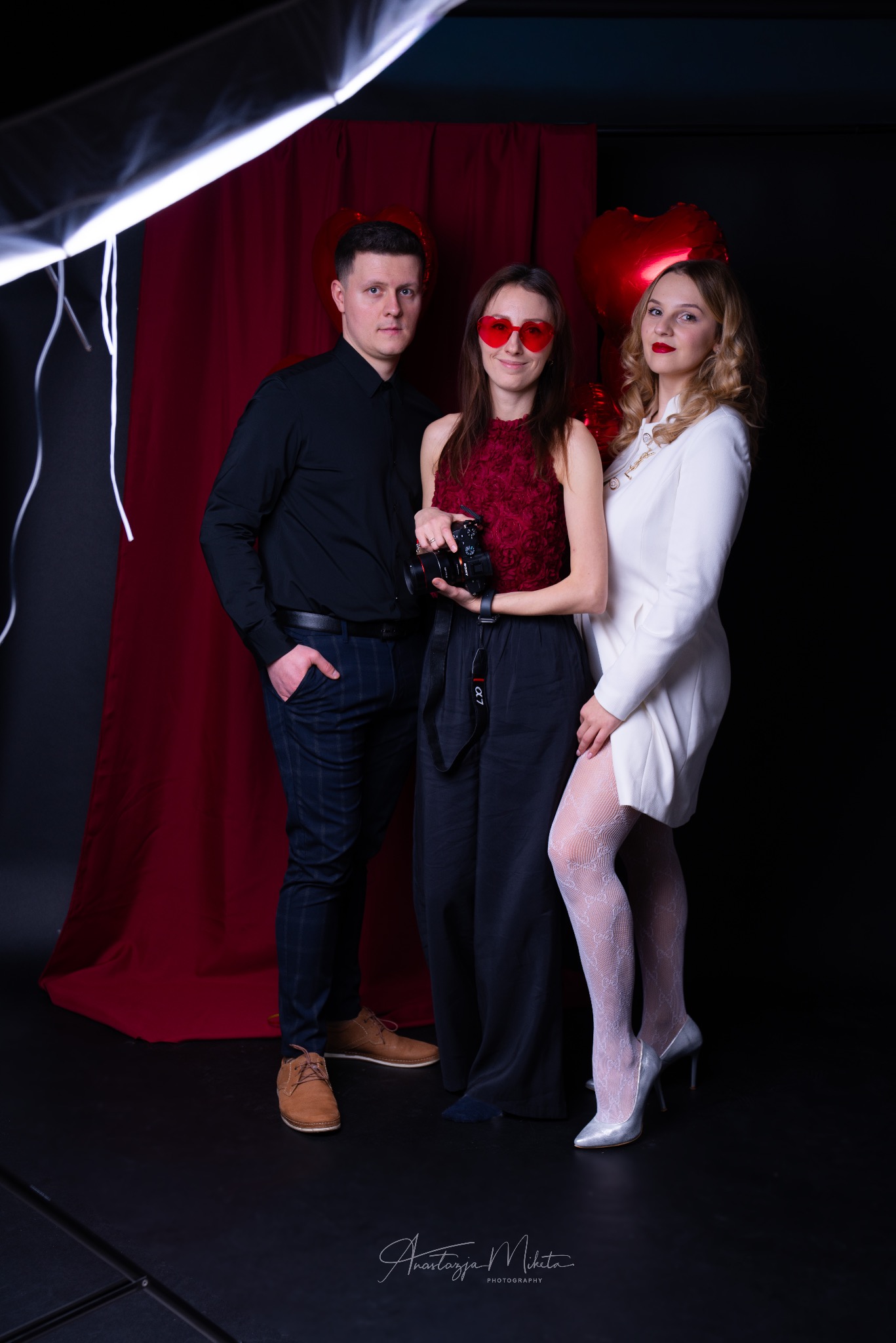 Trzy osoby pozują na tle czerwonej zasłony i balonów w kształcie serc. Kobieta w środku trzyma aparat. Studio fotograficzne z oświetleniem. Sesja w walentynkowym klimacie.